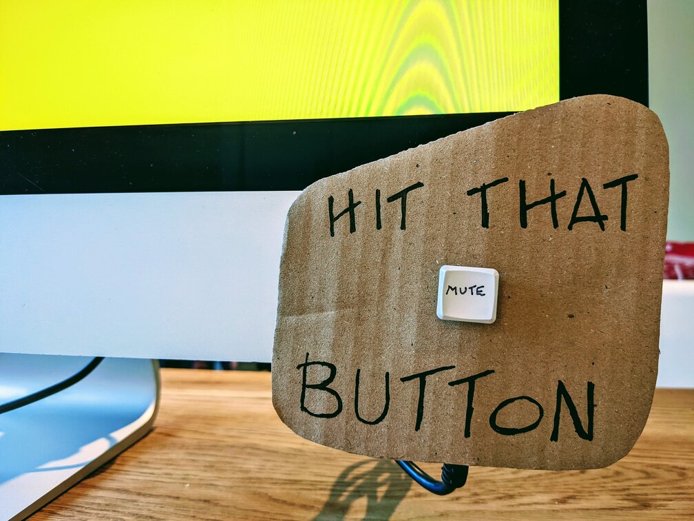 Cardboard sign stuck to a computer monitor with the words "HIT THAT MUTE BUTTON" - the mute button is a computer keyboard button